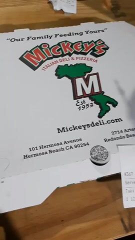 A person opens a pizza box from Mickey’s Italian Deli & Pizzeria to reveal a partially eaten pizza. The pizza is topped with cheese, pepperoni, sausage, tomato sauce, and fresh basil. The pizza box is sitting on a wooden surface, and a receipt is nearby.