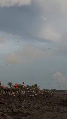The scene captures a seaside view with a predominantly cloudy sky above. Numerous kites are visible at various heights, suggesting kite-flying activities. The ground is covered with large, dark rocks. A cluster of structures, including buildings with orange roofs, is visible in the background along the shore. People are gathered around these structures. The sky is blue with some clouds. A statue is visible amidst the buildings. Overall, the video portrays a coastal setting with a kite-flying event, attracting a crowd of people.