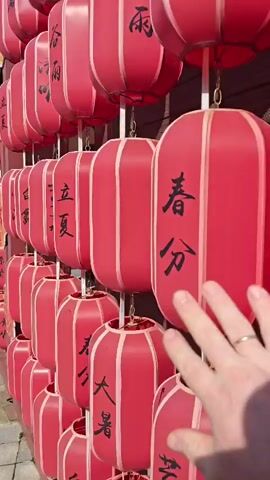The video shows a close-up of a wall adorned with numerous red, cylindrical lanterns. These lanterns are hanging in neat rows, attached to vertical poles with chains. Black Chinese characters are inscribed on many of the lanterns. A person's hand, with a ring on one finger, is seen reaching out and touching some of the lanterns and an orange flower-shaped decoration. The background suggests an outdoor setting with wooden elements and a sunny ambiance. The camera pans slightly, offering different angles of the lanterns and the hand's interaction with them.
