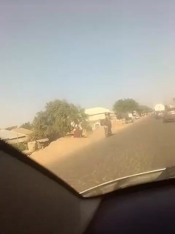 The video, filmed from inside a car, shows a road with a motorcycle rider and several vehicles. A building is visible to the left, along with some trees and sand. The sky is clear and hazy. The car is moving forward on the road. The road is a bit cracked and dusty.  There is light traffic.