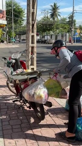 A person wearing a helmet and face covering is standing next to a motorcycle loaded with bags of produce. The motorcycle is parked on a brick sidewalk next to a street. The person is reaching for a plastic bag filled with green produce. Other bags of produce are also visible on the motorcycle and on the sidewalk. In the background, cars and motorcycles are driving on the street. The sky is blue with some clouds. The sun is shining.