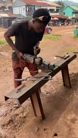 A young man is grinding a piece of metal on a workbench in an outdoor setting. He is wearing a dark t-shirt, patterned red pants, and sandals. Sunglasses are perched on top of his head. The workbench is made of steel and is equipped with a vise. Sparks fly as the man uses an electric angle grinder on the metal clamped in the vise. Other tools and metal pieces are scattered on the workbench. The background shows a dirt road with buildings and market stalls. The ground appears to be damp.