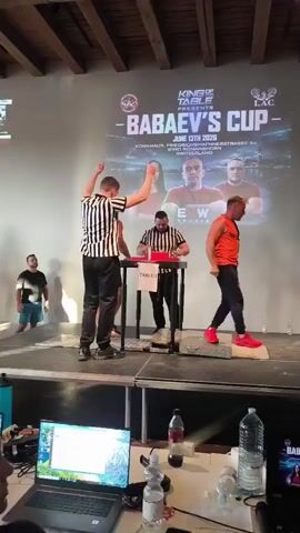 A group of people are gathered on a stage for an arm wrestling competition. The stage is set up with an arm wrestling table in the center, where two men are about to compete. Two referees, dressed in black and white striped shirts, are overseeing the match. A large screen in the background displays the event's logo, "Babaev's Cup," and details about the competition, including the date and location. The audience appears to be seated below the stage, with some spectators visible in the foreground, looking at laptops. The environment suggests an indoor venue with wooden ceiling beams and bright lighting.