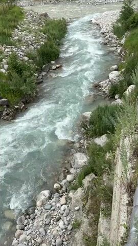 This is a high-angle shot of a turbulent, fast-flowing river. The water is a milky turquoise color and is filled with white foam and rapids. The riverbanks are lined with smooth, grey stones and sparse green vegetation. The shot is taken from a slightly elevated position, looking down the length of the river as it curves to the left and disappears into the distance. There are no people or animals visible in the frame. The overall impression is one of a wild and untamed natural environment.