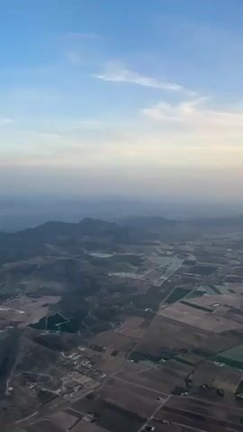 This is an aerial video footage of a rural landscape, likely Elx (Elche), Spain, taken during what appears to be dawn or dusk. The time of day is suggested by the soft, diffused light and the gradient of colors in the sky, transitioning from a clear blue at the top to lighter shades of yellow and white near the horizon, with a hint of haze.

The scene is dominated by a vast expanse of cultivated fields, characterized by their geometric patterns and varying shades of brown and green. These fields are interspersed with patches of darker, possibly wooded areas, and occasional bodies of water that appear as reflective surfaces. Rolling hills and distant mountains form a layered backdrop, diminishing in clarity due to atmospheric perspective, suggesting a wide and open terrain.

Scattered throughout the landscape are small clusters of buildings, indicating human settlement within this agricultural region. The overall impression is one of agricultural activity and a serene, natural environment. There are no visible people or specific objects that stand out, the focus being on the sweeping view of the land. The weather appears to be clear, with a few wispy clouds present in the sky. The mood is tranquil and expansive, conveying a sense of peace and the beauty of the countryside from a bird's-eye perspective. No visible text is present in the footage.