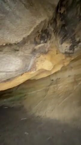 The video pans across the interior of a natural rock shelter or overhang. The rock formations appear to be sandstone, with various layers and textures visible. Darker mineral staining is present on the ceiling of the shelter. The ground within the shelter is sandy and uneven. Towards the right side of the frame, the opening of the shelter is visible, revealing trees and greenery outside. The lighting suggests it is daytime.