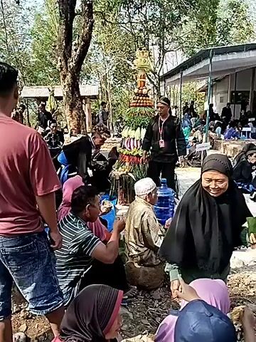 The video shows a gathering of people outdoors near a building with a traditional Indonesian architectural style. Many individuals are wearing black clothing and some have red sashes. Some are seated on the ground, while others are standing or walking. There appears to be some form of ceremony or event taking place, possibly related to a shrine or grave site, as indicated by the presence of what seem to be tombs or grave markers. In the foreground, a woman in a black hijab is carrying a tray. A man in traditional attire is standing and observing the scene, and there are also displays of fruit and other items. The environment suggests a rural or semi-rural setting with trees and a clear sky.