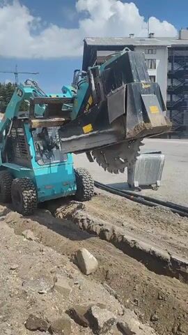 A teal-colored skid steer loader with a trencher attachment is digging a trench in a dirt area. The trencher has a large circular blade that rotates, cutting into the ground. The skid steer is moving forward, and the trencher is creating a narrow, deep trench. There are several large rocks and some debris in the trench. In the background, there is a modern building with balconies and windows, a crane, and trees. The sky is blue with some white clouds, suggesting it is daytime with fair weather. The scene appears to be a construction site, possibly for utility installation. There is text visible on the trencher attachment, including "ALPINA" and some yellow warning labels. No people are clearly visible in the frame. The location context indicates Bursa, Turkey, but there are no specific visual cues in the image to confirm this.