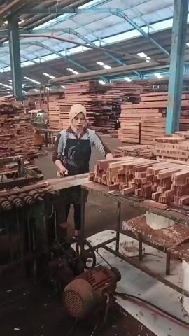 A woman in a headscarf and apron works in a wood processing facility. She is standing in front of a machine and a table laden with wooden pieces. Stacks of lumber are visible throughout the warehouse.
