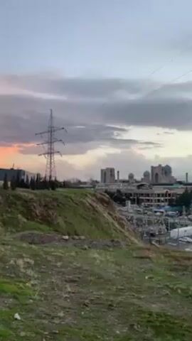 The video showcases a cityscape from an elevated viewpoint, capturing a blend of urban infrastructure and natural elements. In the foreground, a grassy hillside leads down to a parking area filled with vehicles, some painted with graffiti. Overhead, power lines stretch across the sky, supported by a towering electrical pylon to the left of the frame. The background reveals a skyline punctuated by buildings with distinct blue domes, suggesting religious architecture. Beyond the immediate buildings, a highway overpass carries traffic, with cars and trucks visible moving along it. The sky is filled with clouds, tinted with the warm hues of a setting sun, creating a dramatic backdrop to the urban scene. The overall impression is a snapshot of a city on the cusp of dusk, with its mix of industrial, residential, and religious elements.