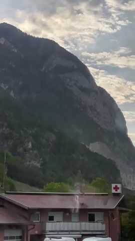 A video captures a view from a moving vehicle, likely a car or bus, through a window. The scene features a large, rugged mountain dominating the background, with a cloudy sky above. In the foreground, a building with a red roof is visible, and on top of it, a red cross emblem is displayed. Trees and greenery surround the building and extend up the lower slopes of the mountain. The camera perspective suggests motion, with the landscape appearing to pass by. There are no discernible people in the frame, and the primary focus is on the natural landscape and the building with the red cross. The lighting indicates it might be daytime, possibly early morning or late afternoon, given the softness of the light.