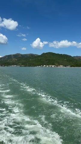 The video captures a scenic view from a moving boat on a bright, sunny day. The turquoise water is choppy, leaving a white foamy wake trailing behind the vessel. In the distance, a lush green, mountainous coastline stretches across the horizon. A small town or village is visible nestled along the shoreline, with buildings peeking out from among the trees. The sky above is a vibrant blue, dotted with fluffy white clouds. The overall impression is one of a peaceful and picturesque journey, likely a tourist excursion or a mode of transport across the water. The time of day appears to be daytime, given the bright sunlight and clear sky. The location is identified as Nam Ninh Hòa, Vietnam, suggesting a coastal or bay area. There is no visible text in the video.