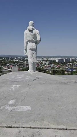 A video pans slowly across a white statue of a person from the back, standing on a concrete platform. The statue faces away from the camera, looking out over a city with houses and buildings, and a clear blue sky.