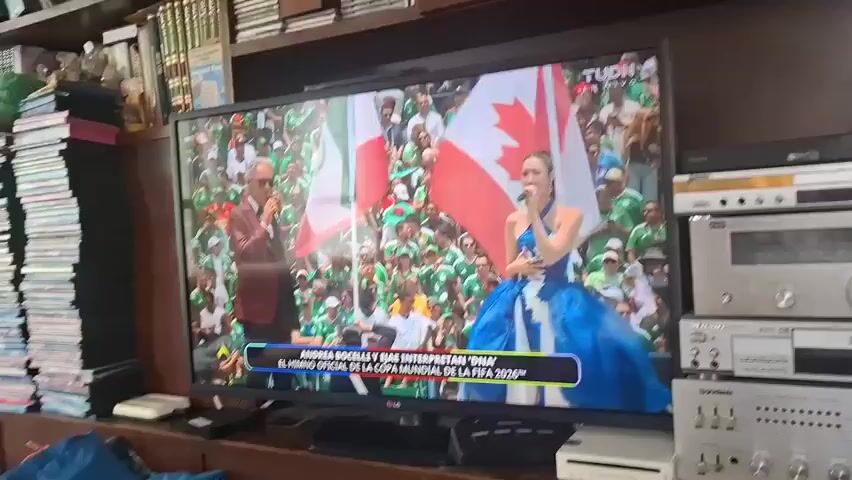 The video shows a television screen displaying a performance. On the screen, a woman in a blue dress is singing, holding a microphone. Beside her, a man in a suit is also present. Behind them, a crowd of people are visible, many of them wearing green. Two large flags, one red and white and the other red and white with an emblem, are displayed prominently. A banner at the bottom of the screen reads "ANDREA BOCELLI Y EIAE INTERPRETAN 'DINA' - EL HIMNO OFICIAL DE LA COPA MUNDIAL DE LA FIFA 2026". The television is placed on a wooden surface. To the right of the television, there are several electronic devices stacked vertically, including what appears to be a VCR and an audio receiver. To the left of the television, a stack of books or DVDs is visible. The background suggests an indoor setting, possibly a living room or entertainment area. The lighting is somewhat dim.