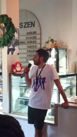 The video shows a person standing in a cafe, holding a microphone and speaking. The person has a beard and is wearing a white t-shirt with a purple graphic, black shorts, and a lanyard around their neck. They are leaning on a counter with a display case behind them filled with baked goods. Above the display case is a sign that reads "SZEN" with a list of prices. There's also a red doll and a wreath. In the background, another person walks behind the counter. The cafe has a bright, modern aesthetic.