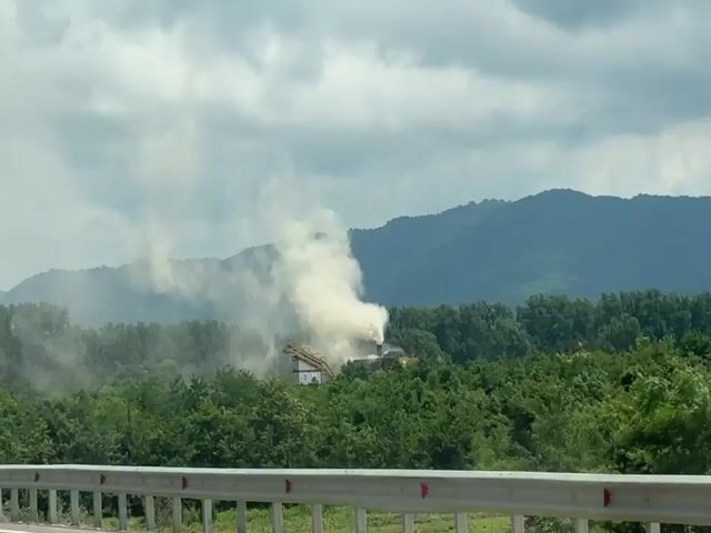 The video shows a rural landscape with a dense forest in the foreground and rolling hills in the background. A plume of white smoke rises from a structure situated within the forest. There are several road signs visible. One sign is green with a white number 24 inside a square. Another sign is white with a red circle containing the number 110, indicating a speed limit, and a white rectangular sign above it with text that appears to be in a foreign language and reads "SECTION SPEED VIDEO CONTROL". The smoke appears to be emanating from an industrial or processing facility. The sky is cloudy, suggesting overcast weather conditions. The camera is positioned behind a guardrail, indicating the footage was likely captured from a moving vehicle on a road overlooking the scene. There are no people or animals visible in the frame.