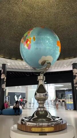 The video shows a large, ornate globe displayed on a polished metal stand in a spacious, modern interior. The globe itself is adorned with a sparkling blue surface and distinctly colored continents. Behind the globe, several people are visible, some sitting on a blue sofa and others standing or walking. In the background, there are displays and storefronts, one of which appears to be for "Swarovski" and features a large winged figure. The ceiling above the globe is dark and speckled with lights, creating a starry effect. The overall environment suggests a public indoor space, possibly a shopping mall or an exhibition hall.