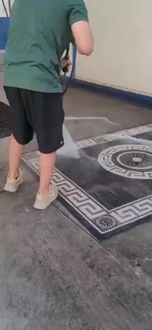 A person wearing a green shirt and black shorts is pressure washing a rug on a concrete surface. The rug is dark with a white and grey geometric pattern. The person is holding a pressure washer wand and spraying water onto the rug, creating a spray of water and foam. The rug is partially wet and soapy. The background appears to be an outdoor or semi-outdoor area with a wall and a blue trim.