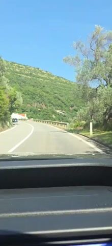 The video captures a POV shot from inside a vehicle driving on a winding road. The road is paved with white lane markings and a wooden guardrail runs along the right side. To the left of the road, there are trees and a steep, green, tree-covered hillside rising upwards. In the distance, a white building with an orange roof is visible nestled on the hillside. The sky is clear and blue. The vehicle appears to be moving at a moderate speed, and the overall environment suggests a rural or mountainous area on a sunny day.
