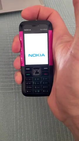 A hand holds a black and pink Nokia XpressMusic cell phone, which is powered on and displaying the Nokia logo followed by a notification in Hungarian: "Az akkufeszültség alacsony" (Battery voltage is low). The phone's screen shows a low battery icon. The phone is held over a textured blue surface, possibly a table or cloth, with the corner of a silver laptop visible in the upper left portion of the frame. The background is blurred.