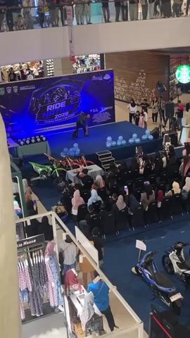 This media file captures an indoor event taking place in a shopping mall, likely in Dumai City, Indonesia, as indicated by the banner text. The scene is set on a blue carpeted floor with a stage at the back. A large, brightly lit banner on the stage reads "RIDE NATION 2023" and features graphics related to motorcycles. A presenter, dressed in dark clothing, stands center stage, seemingly addressing an audience.

In front of the stage, a green dirt bike is displayed. Several rows of chairs are arranged for an audience, with many people, predominantly women in hijabs, seated and facing the stage. Some audience members are holding up their phones, possibly recording or taking pictures. To the right of the stage, a group of children and adults stand, observing the event.

Below the stage area, there are displays of clothing, with mannequins and racks of garments visible. Further to the right, two scooters are parked. The lighting suggests it is daytime, and the environment is clearly indoors within a commercial space, evidenced by the tiered levels of the mall and other storefronts in the background, including a Starbucks. The overall atmosphere appears to be one of an organized public event, possibly a product launch or exhibition related to motorcycles, with an engaged audience.