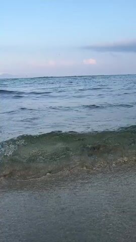 A low-angle shot captures the gentle lapping of clear water on a sandy beach. The sun is low in the sky, casting a soft light on the scene. Small waves roll in from the ocean, creating a soothing rhythm. The water is shallow, allowing the sandy bottom to be visible. The sky is a soft blue with a few wispy clouds. The overall atmosphere is peaceful and serene.