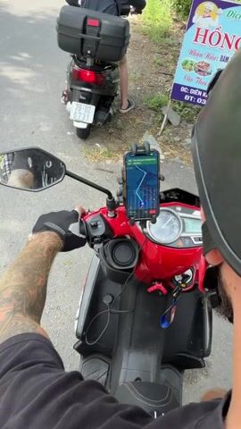 The video shows two people on motorcycles on a paved road next to vegetation and trees. In the foreground, a person wearing a black glove on their left hand is on a red and black motorcycle. A smartphone is mounted to the handlebars, displaying a navigation app with a route. The person's tattooed arm is visible. In the middle ground, a person wearing a helmet and a black hoodie is on a black motorcycle with a black storage box on the back. A pink car is parked on the side of the road to the left. A sign with Vietnamese text is visible on the right side of the road. The video appears to be filmed from the perspective of the person on the red motorcycle, following the person on the black motorcycle.