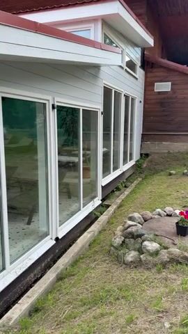 The video shows a building with large windows and a red roof. The exterior walls are white, with grey siding at the bottom. There is a gutter system visible on the roof, with a downspout running down the side of the building. The ground around the building is covered in grass, with some rocks and a wooden walkway. The interior of the building is visible through the windows, showing furniture and what appears to be a tiled floor. The sky is cloudy with patches of blue. Trees are visible in the background. The camera is moving, panning across the exterior of the building and its surroundings.
