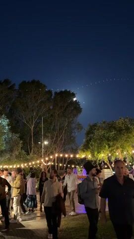 This full-length, vertical shot captures an outdoor nighttime gathering in Nir Eliyahu, Israel. Numerous people, mostly adults, are seen mingling, walking, and conversing in a lively social atmosphere. The scene is illuminated by warm string lights draped across trees and overhead, casting a festive glow. The ground alternates between paved areas and grassy sections.

In the background, lush, leafy trees frame the event against a dark blue night sky. A strikingly bright, almost full moon dominates the upper mid-frame, shining through the branches of the trees. Above the moon, a distinct line of small, bright points of light stretches across the sky, characteristic of Starlink satellites.

People are dressed in casual attire, with some individuals wearing hats. In the mid-ground, to the right, a person stands behind what appears to be a bar or a service station. The overall mood seems relaxed and enjoyable, with clear skies and ample lighting contributing to a pleasant evening event. There is no visible text within the image.