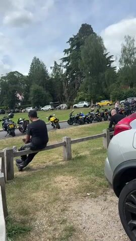 The video shows a gathering of people and motorcycles in a grassy, outdoor setting on a partly cloudy day. There are numerous motorcycles parked in a row on a paved area that appears to be a street or parking space. Several cars are visible in the background as well. A man in a black shirt and motorcycle gear is sitting on a wooden fence, seemingly observing the scene. A green trash can is next to him. As the camera pans to the left, more motorcycles come into view, lined up neatly. People are seen standing and talking near a small green building or shed. The area is surrounded by trees and buildings, creating a village or town atmosphere. A large stone pillar is also visible in the background.