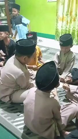 A group of young boys, all wearing peci caps, are seated on the floor in a room. Some are wearing traditional Muslim clothing, while others are in more casual attire. They appear to be engaged in a game or activity, with some of them looking at each other and smiling. The room has green walls and a patterned curtain in the background.