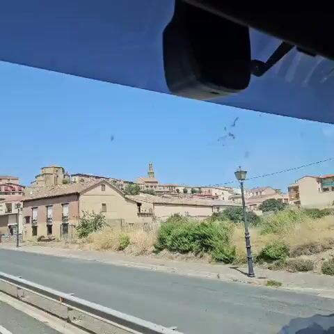 A video shows a view from a vehicle traveling along a road. On the right side of the road, there are buildings with brick and stucco facades, some with red-tiled roofs and balconies. Trees with dark purple foliage line the street. In the background, a town is visible on a hill, with several buildings, including a prominent domed structure and a tower. The sky is clear and blue. The video appears to be filmed on a sunny day. There are some spots on the window, possibly dirt or water marks.