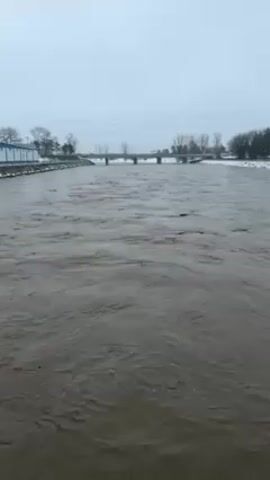 The video shows a muddy river flowing rapidly under a bridge in an overcast, wintery scene. Snow is visible on the banks of the river. The water is turbulent, suggesting a high flow rate. The bridge appears to be a standard, multi-span structure. The sky is a uniform gray, indicating a cloudy or overcast day. The presence of trees without leaves suggests it is late fall or winter. The riverbanks are lined with vegetation or structures, some of which are painted blue and white.