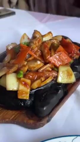 The scene is a close-up shot of a sizzling dish on a white tablecloth. The dish contains meat, mushrooms, onions, and carrots. The food is covered in a dark, glossy sauce, and the sizzling sound suggests it is being cooked on a hot stone or metal plate. The plate is a dark color, and sits on a wooden tray. The table around the dish is covered with a white tablecloth. The overall impression is of a freshly prepared and appealing meal.