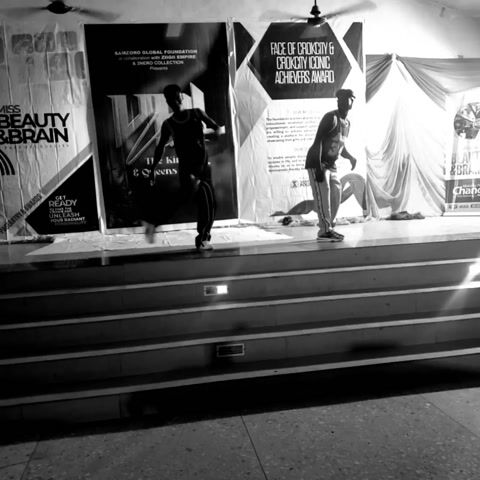 In black and white, two figures dance on a stage with several posters behind them. The stage is elevated with steps leading up to it, each step illuminated by recessed lights. One figure is dressed in dark clothing, while the other wears a basketball jersey. Their movements are energetic and coordinated, suggestive of a choreographed performance. The setting appears to be an event or competition, with banners promoting awards and foundations.