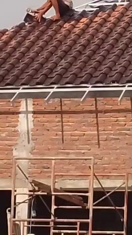 A man is working on a tiled roof. He is sitting on the roof and appears to be placing or adjusting tiles. There is scaffolding and a brick wall below the roof. The sky is overcast.