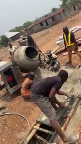 The scene depicts a construction site with several workers engaged in pouring and leveling concrete. In the foreground, a man in a black shirt and gray shorts is using a tool to smooth the concrete within a wooden framework. Behind him, a worker in an orange vest is seated on a pile of broken concrete, observing the work. A large cement mixer is pouring its contents onto the broken concrete, where another worker is managing the flow. To the right, a man wearing a yellow safety vest is standing, seemingly supervising the operation. The backdrop reveals a rural setting with simple buildings and sparse vegetation under a bright sky. The overall environment is dusty, and the ground is predominantly red soil.