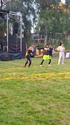 This is a vertical video taken outdoors during the daytime. The scene is a grassy field in front of a raised stage with a black backdrop and a lighting rig. Several people are dancing on the grass. In the foreground, a person in a yellow and black outfit is dancing, facing away from the camera. To their right, another person in a black outfit with colorful leggings is dancing. Further to the right, a person in a neon green and black outfit is dancing. In the background, on the stage, several individuals are visible, some appearing to be part of a performance or setup. The overall atmosphere appears to be an outdoor event or performance.