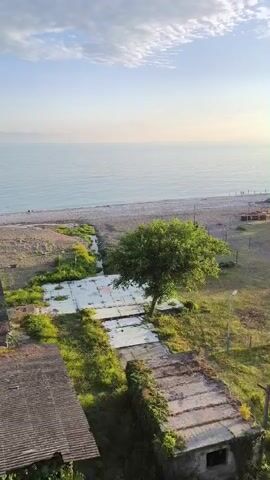 The video begins with an aerial view of a beach and a body of water under a clear sky with some clouds. The scene transitions to show an area with grassy fields, some overgrown structures, a lone tree, and a playground. Further along, a fenced sports field is visible with several people playing a game, likely soccer, under the setting sun. The sun is bright and casts long shadows across the field. Buildings and more trees are visible in the background, along with a glimpse of the sea.