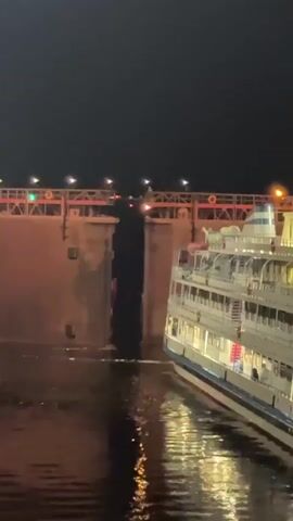 A large white cruise ship with multiple decks and illuminated windows is positioned beside a tall, concrete lock gate structure at night. The lock gate has red and green lights along its top edge. The ship's bow is pointed towards an opening in the lock gate. Water is visible in the foreground, reflecting the lights from the ship and the lock. The scene is dark, suggesting it is nighttime or twilight.