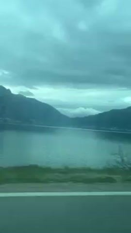 The video shows the view from a moving vehicle, likely a car. The landscape consists of a large body of water, possibly a lake or bay, with mountains rising in the background. The sky is overcast with a uniform layer of clouds. In the foreground, there are bare trees along the roadside, indicating it may be a colder season. The road itself is visible, with a white line marking the edge. The overall color palette is muted and dominated by shades of blue and green. The camera appears to be handheld or mounted inside the vehicle. The focus varies as the vehicle moves, occasionally blurring the foreground elements.