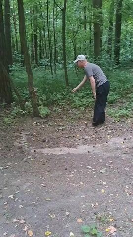 This media file shows a man in a forest. He is wearing a grey t-shirt, black pants, and a white baseball cap. The man is bending down, reaching out towards the ground with his right hand. He appears to be examining or interacting with something on the forest floor.

The scene is a wooded area with many trees and dense foliage. The ground is covered with dirt, fallen leaves, and small debris, possibly pine cones or twigs. There's a path or clearing in the foreground, and the background is filled with the trees and undergrowth characteristic of a forest. The lighting suggests it is daytime, possibly overcast or in a shaded part of the forest, as there are no harsh shadows. The overall atmosphere seems calm and natural.

The man's posture suggests focus and a gentle interaction with his surroundings. There are no visible indications of specific weather conditions beyond the general appearance of a humid forest environment. The location is identified as Lytkarino, Russia. There is no visible text in the media file.