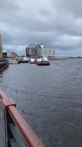 The video shows a body of water with several boats. On the left side of the frame, there is a wooden railing with a dark red top. In the background, there are buildings and a cloudy sky. The water is choppy, indicating wind or current. The overall scene is muted, with a focus on the overcast weather and the industrial/urban waterfront.