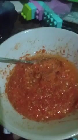 A close-up shot of a bowl of red chili sauce with a spoon in it. The sauce appears to be freshly made, with visible chili seeds and liquid. In the background, there are some colorful building blocks and a purple plastic bag.