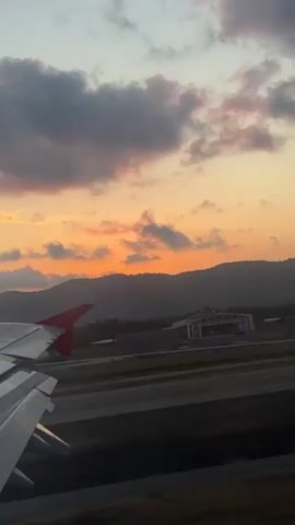 A long vertical shot, from a window seat on an airplane, captures the sunset and a red-tipped wing of the plane while it is on a runway. A brilliant orange sun descends over the silhouette of a mountain range. Above the mountains are large, dark, fluffy clouds, some of which are tinged with pink. In the foreground, below the airplane wing and to the left, is a runway. To the right, below the mountain range, are several buildings, the top of a control tower, and other facilities, suggesting an airport. The airplane appears to be either landing or taking off due to its presence on the runway and the visible landscape. The sky is a mix of blues, oranges, pinks, and grays.