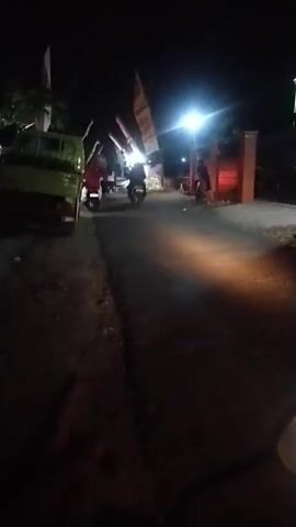 The video shows a nighttime scene on a paved road. Several motorcycles are visible, some moving and some parked. One green truck is parked on the left side of the road. There are flags or banners on poles lining the right side of the road, illuminated by a bright street light. The road is dark, with some areas illuminated by the headlights and taillights of the motorcycles. The overall impression is of a somewhat rural or suburban street at night, possibly in a place where nighttime activities or gatherings are taking place. There are a few blurry figures on the motorcycles, but their identities are not discernible. The video appears to be shot from the perspective of someone riding a motorcycle, as indicated by the partial view of a handlebar and headlight in the foreground of the final frames. The lighting is dim, and there is some motion blur, suggesting movement during the recording.