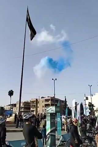 The video shows a Syrian flag on a tall flagpole against a blue sky with scattered white clouds. To the right of the flag, a cloud of bright blue smoke is visible. Below the flag, a group of people are gathered on what appears to be a street or plaza. Many of them are wearing helmets, and some are holding or riding bicycles. There are also several banners and signs with Arabic text visible within the group. The background includes buildings, streetlights, and palm trees. The overall scene suggests a public event or gathering taking place in Syria.