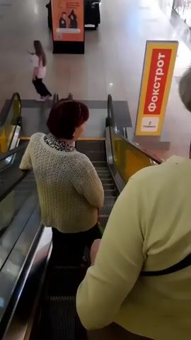 Two women are on an escalator in what appears to be a shopping mall. The woman in the foreground is wearing a light green shirt and is partially visible from behind. The woman in the middle is wearing a white knitted sweater and a dark skirt. She is facing away from the camera and looking to her right. An advertisement sign for "ФОКСТРОТ" is visible on the right side of the frame. The escalator is moving downwards.