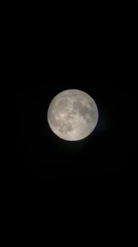 A full moon hangs in the black sky, slightly to the left of the center of the frame. The moon is illuminated, revealing craters and maria on its surface. There are faint lens flares visible at the bottom right of the moon, one green and one blueish. The background is entirely black.