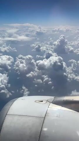 A view from an airplane window shows a vast expanse of white and gray clouds under a clear blue sky. The edge of an airplane engine nacelle, made of segmented metallic panels, occupies the bottom portion of the frame. The nacelle is a dull silver color and shows some signs of wear and tear, with visible rivets and seams. The clouds are dense and puffy, with some appearing darker and heavier than others, suggesting varying altitudes and thicknesses. Sunlight glints off the metallic surface of the nacelle, creating subtle highlights. The overall scene conveys a sense of altitude and travel, with the clouds forming a textured, layered landscape beneath the aircraft.